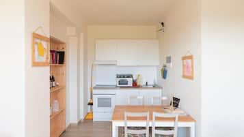 Cocina compacta con armarios blancos, microondas y horno pequeño. Hay una mesa de madera con sillas y un estante con libros y botellas.
