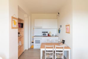 A compact kitchen with white cabinets, a microwave, and a small oven. There is a wooden table with chairs and a small shelf with books and bottles.