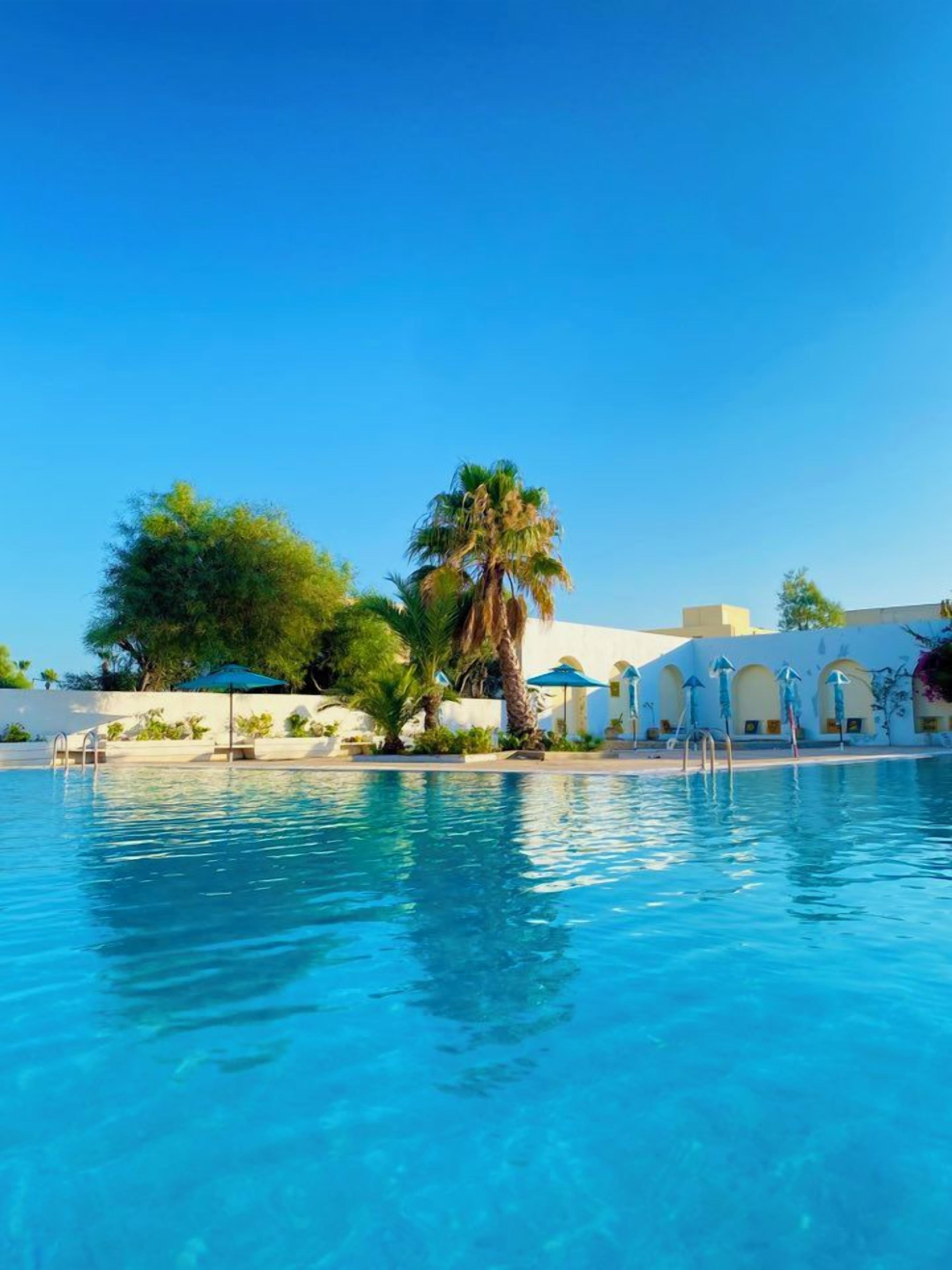 A swimming pool with clear blue water, surrounded by palm trees and a building with arches.