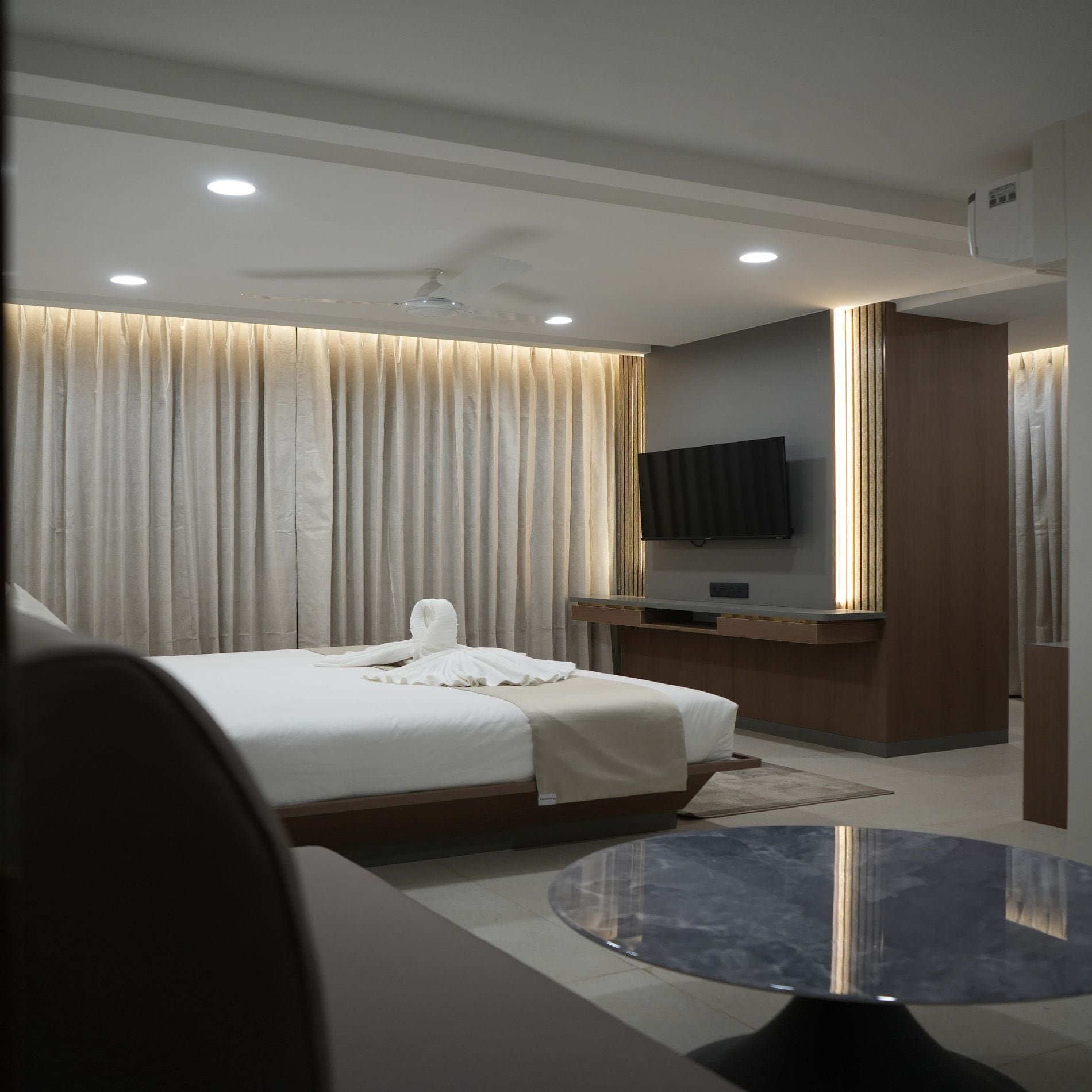 A modern hotel room with a bed, a flat-screen TV, a round table, and a large window with curtains.