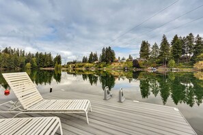 A wooden deck with lounge chairs overlooking a calm lake, surrounded by trees and buildings. - Dock & Views! Waterfront Lake Tapps Retreat (Lake Tapps)
