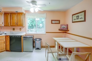 A kitchen with wooden cabinets, a dishwasher, a refrigerator, a trash can, and a window with a view of greenery.