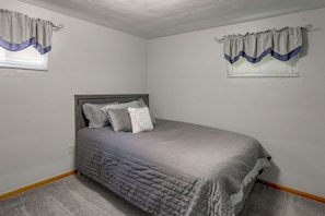 A bedroom with a single bed, a headboard, and two windows with curtains.