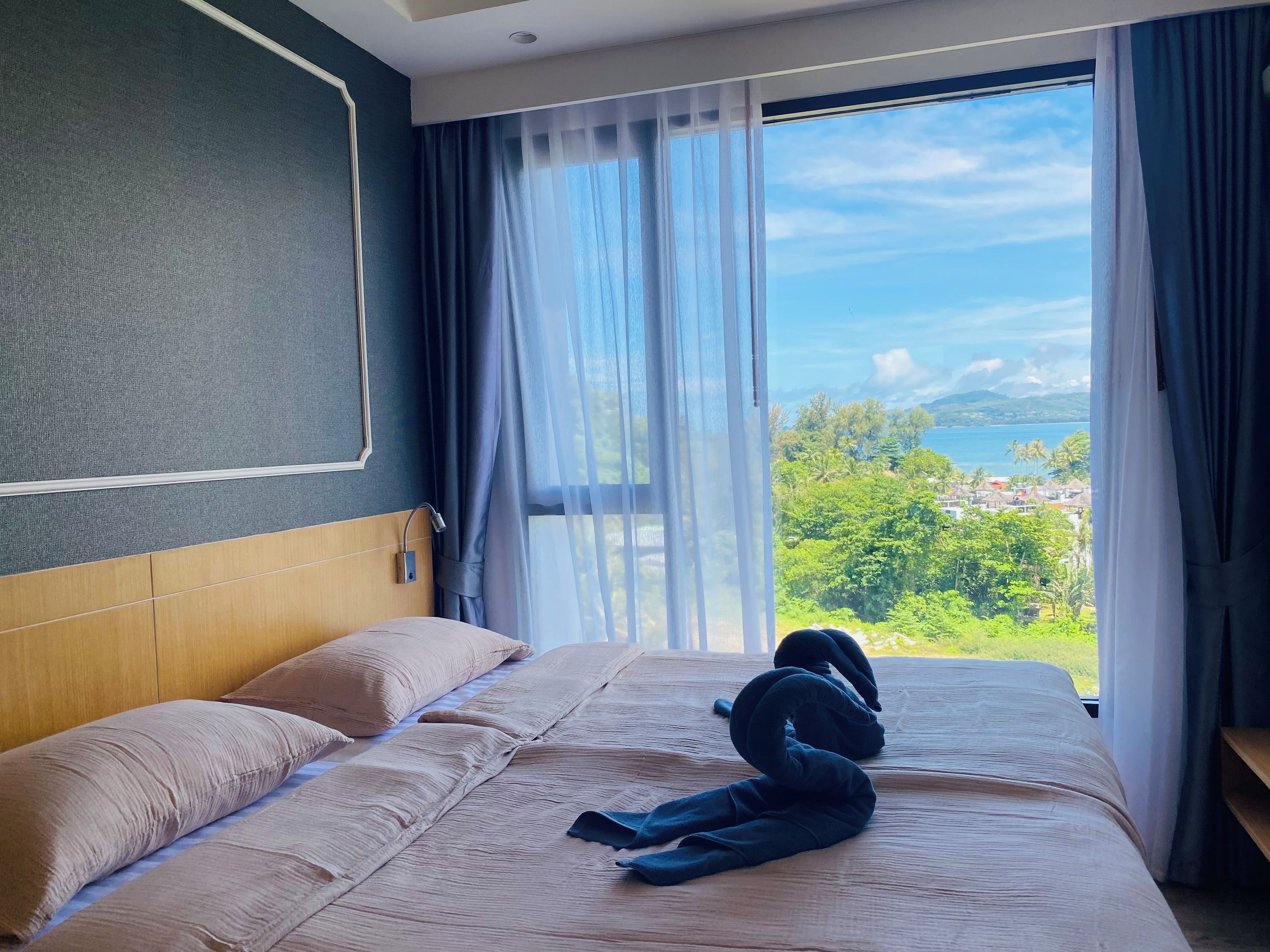 A hotel room with a bed, a view of a coastal landscape, and a window with sheer curtains.
