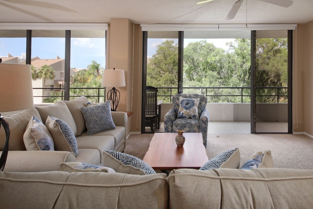 A spacious living room with a beige sofa set, a coffee table, a TV, and a balcony with large windows.
