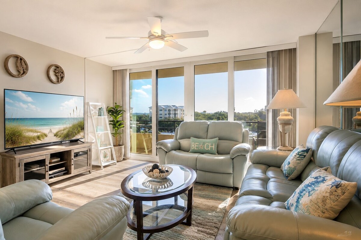 A modern living room with a television, leather sofas, a coffee table, and a balcony with a view of the beach.