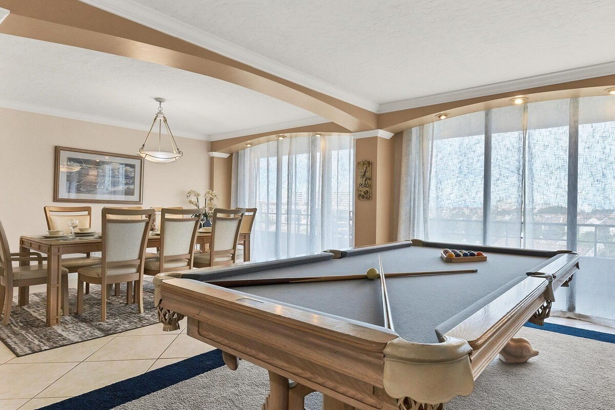 A pool table in a room with a dining area, large windows, and a chandelier.
