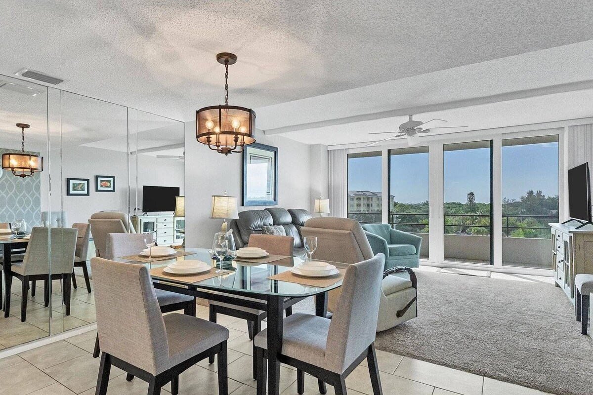 A modern living room with a large glass door leading to a balcony, a dining area with a glass table, and a ceiling fan.