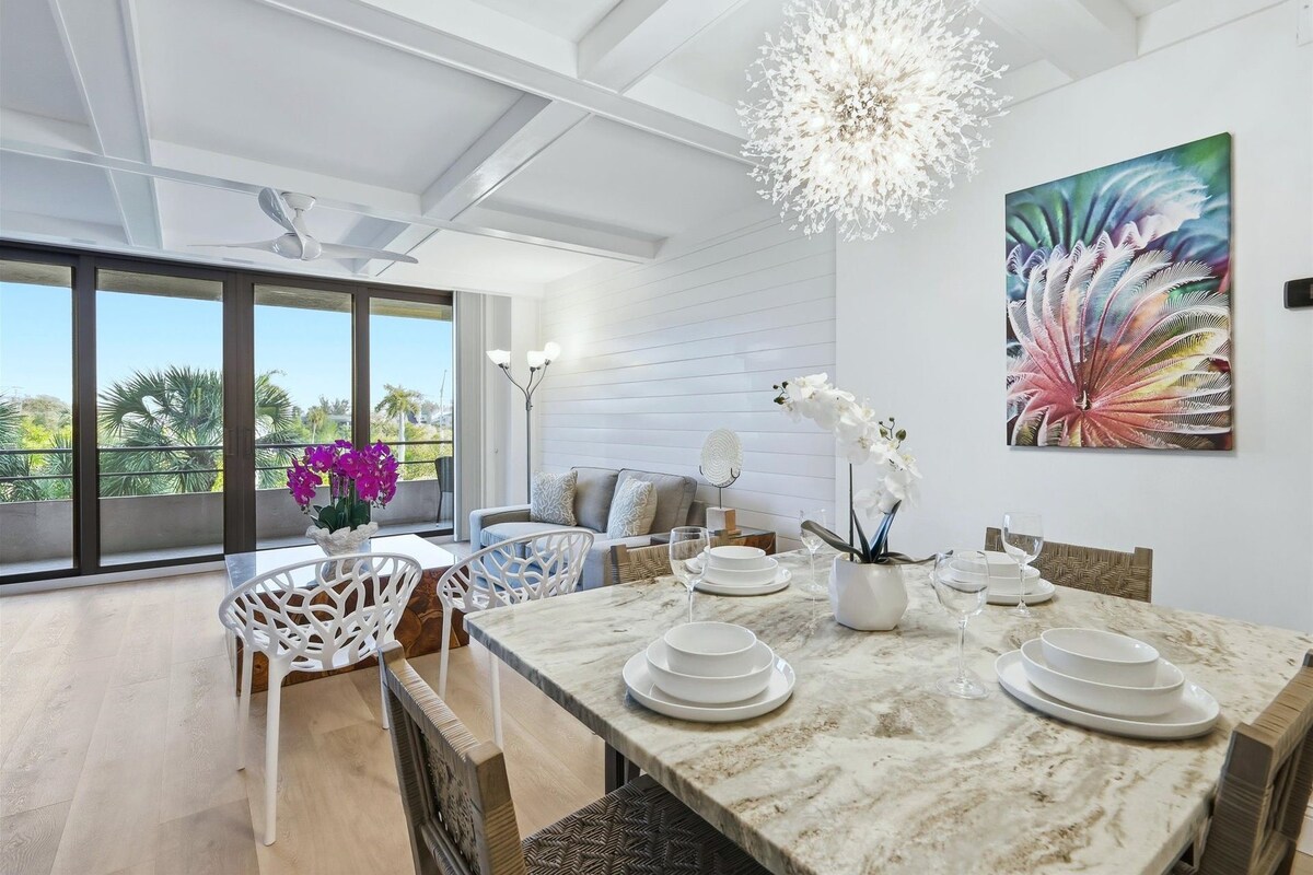 A modern dining area with a marble table, white chairs, and a large window offering a view of palm trees.