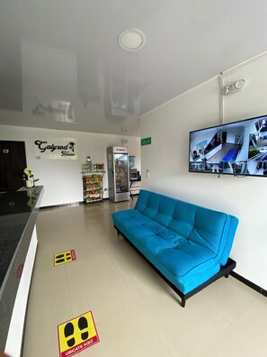 A hotel lobby with a blue sofa, a refrigerator, a water dispenser, and a wall-mounted TV displaying security cameras. - GalyRod House Hotel (San Andrés)