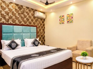 Habitación de hotel con una cama grande, cabecera, sofá, una mesita redonda con una planta y dos obras de arte coloridas en la pared.