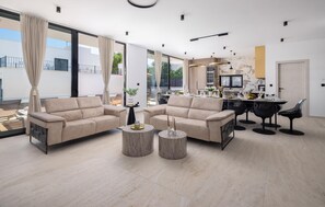 Uma sala de estar moderna com um sofá bege, duas mesinhas de centro e um espaço de jantar com mesa posta para seis pessoas.