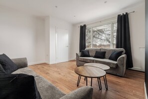 A modern living room with a grey sofa, a round wooden coffee table, and a large window with curtains. - Refurbished Farnworth Home Comfort and Convenience Await for Holiday Makers (Farnworth)