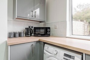 A compact kitchen with a microwave, kettle, and coffee maker on the counter, a washing machine, and cabinets above. - Refurbished Farnworth Home Comfort and Convenience Await for Holiday Makers (Farnworth)