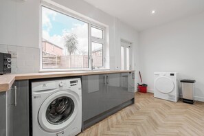 A modern laundry room with a washing machine, a dryer, and a window overlooking a residential area. - Refurbished Farnworth Home Comfort and Convenience Await for Holiday Makers (Farnworth)