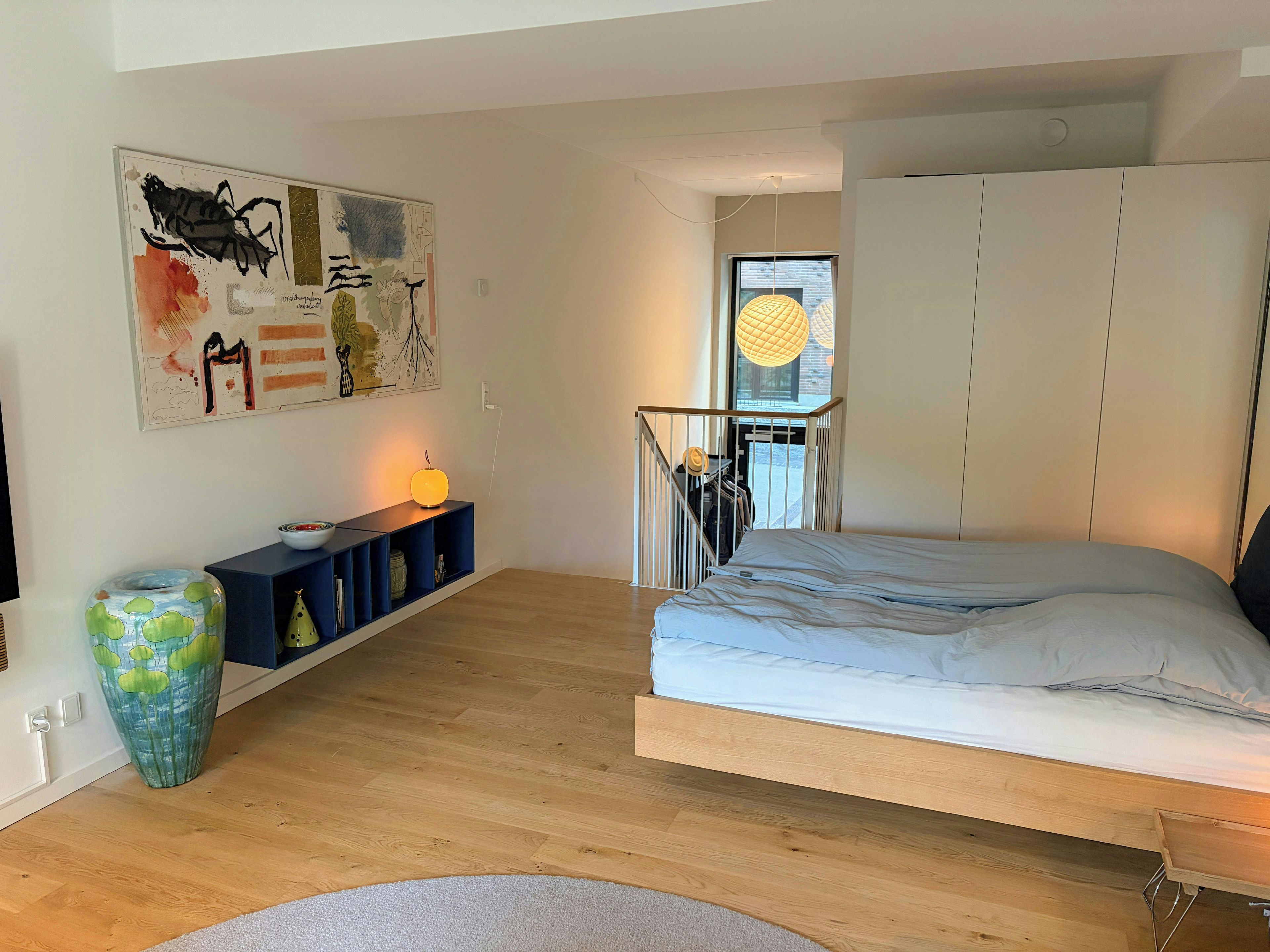 A bedroom with a bed, a nightstand, a lamp, a wall art piece, and a window with a view.