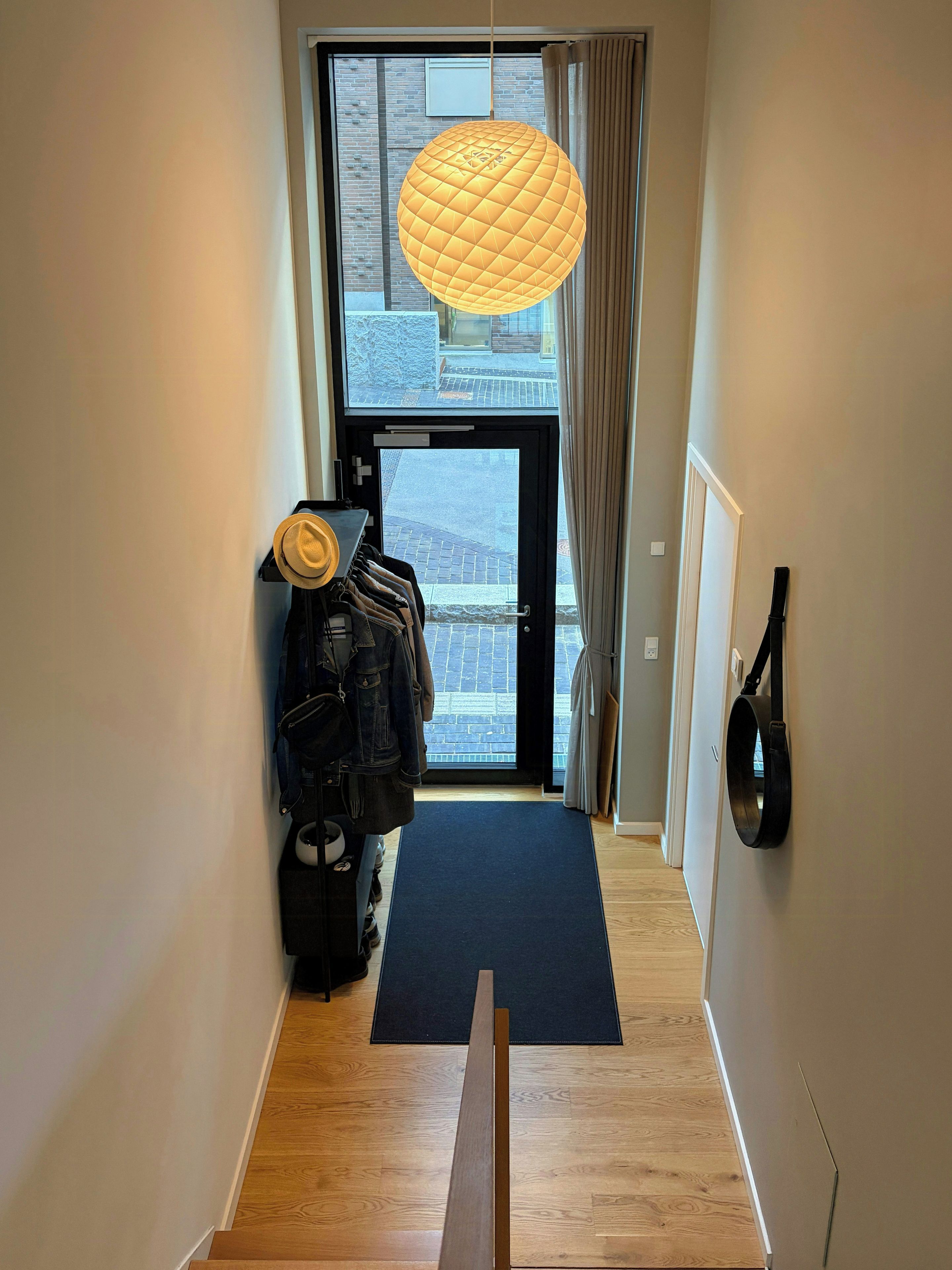 A hallway with a wooden floor, a black mat at the entrance, a hanging light fixture, and a view of the street outside.
