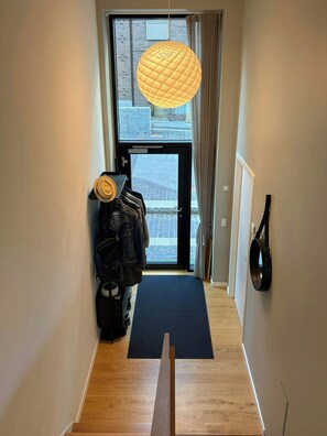 A hallway with a wooden floor, a black mat at the entrance, a hanging light fixture, and a view of the street outside. - Two-storey flat by Carlsberg's garden (København)