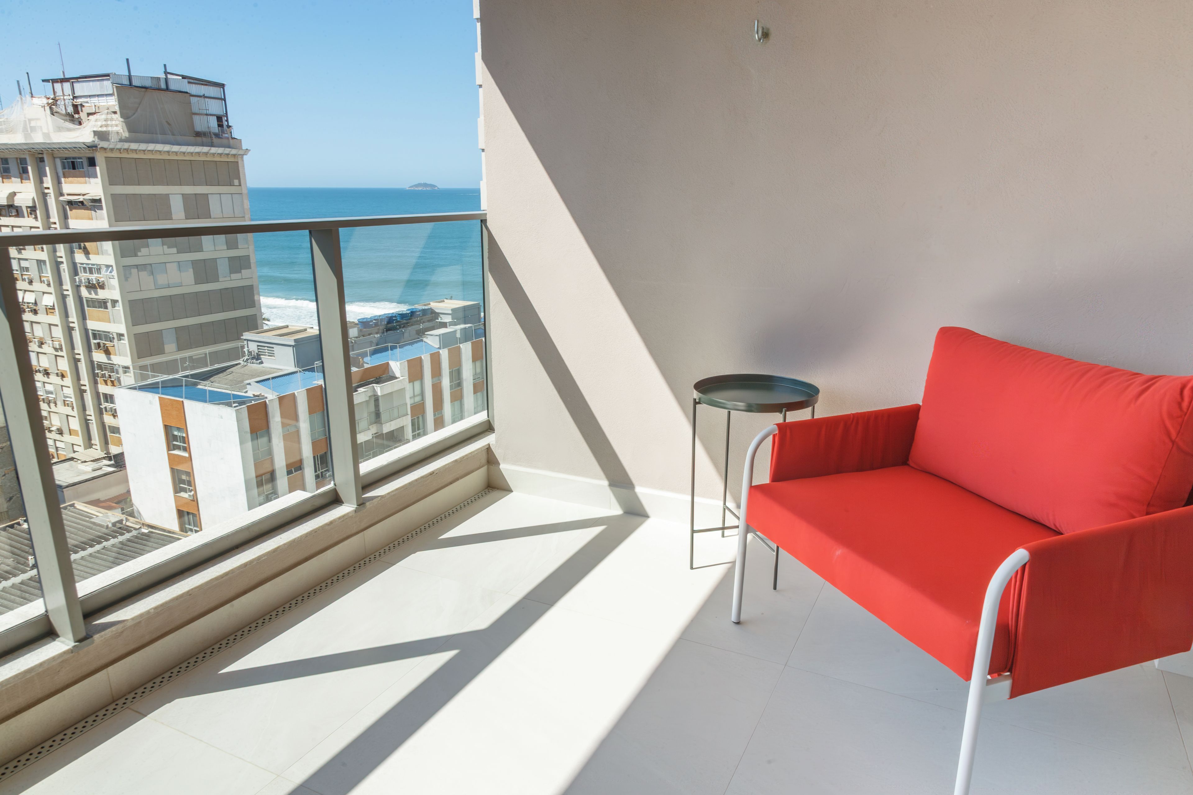 Ein Balkon mit einem roten Stuhl, einem kleinen Tisch und Blick auf das Meer und die Gebäude.