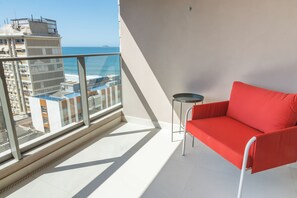 Un balcón con una silla roja, una mesita y vista al mar y edificios.