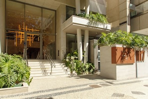 Una entrada moderna de edificio con fachada de vidrio, escaleras que conducen a un balcón y diversos maceteros con plantas.