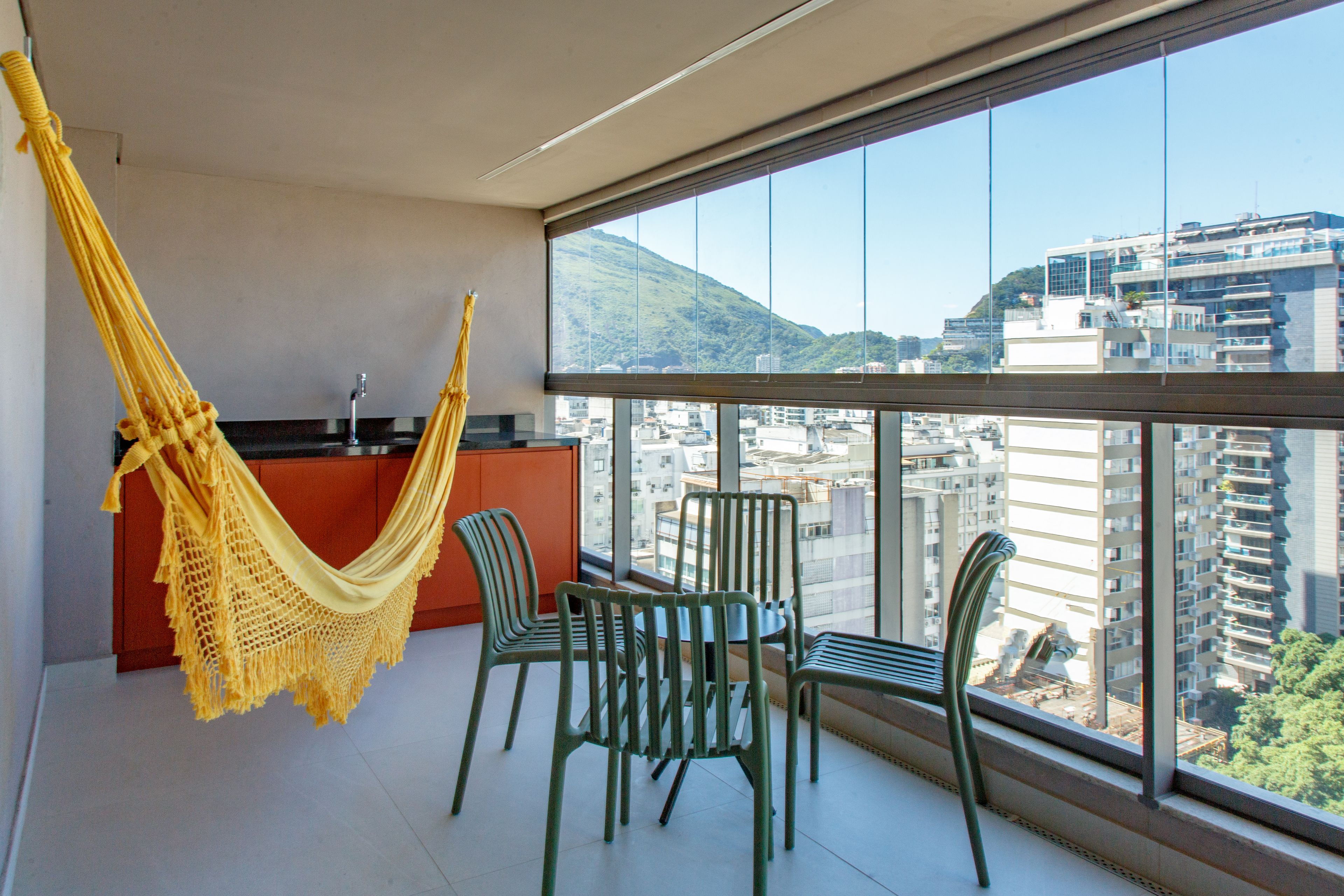 Un balcón con vista a un paisaje urbano, una hamaca amarilla y sillas verdes.