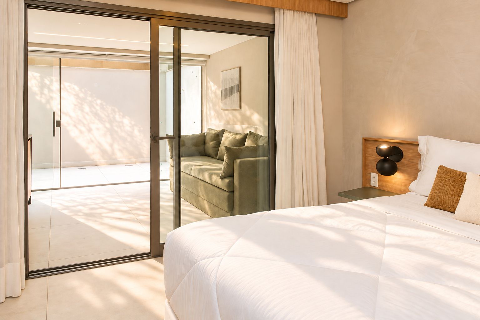 Un dormitorio con una cama grande, una mesita de noche con lámpara, una puerta corrediza de vidrio que da a un balcón y un sofá en la habitación contigua.