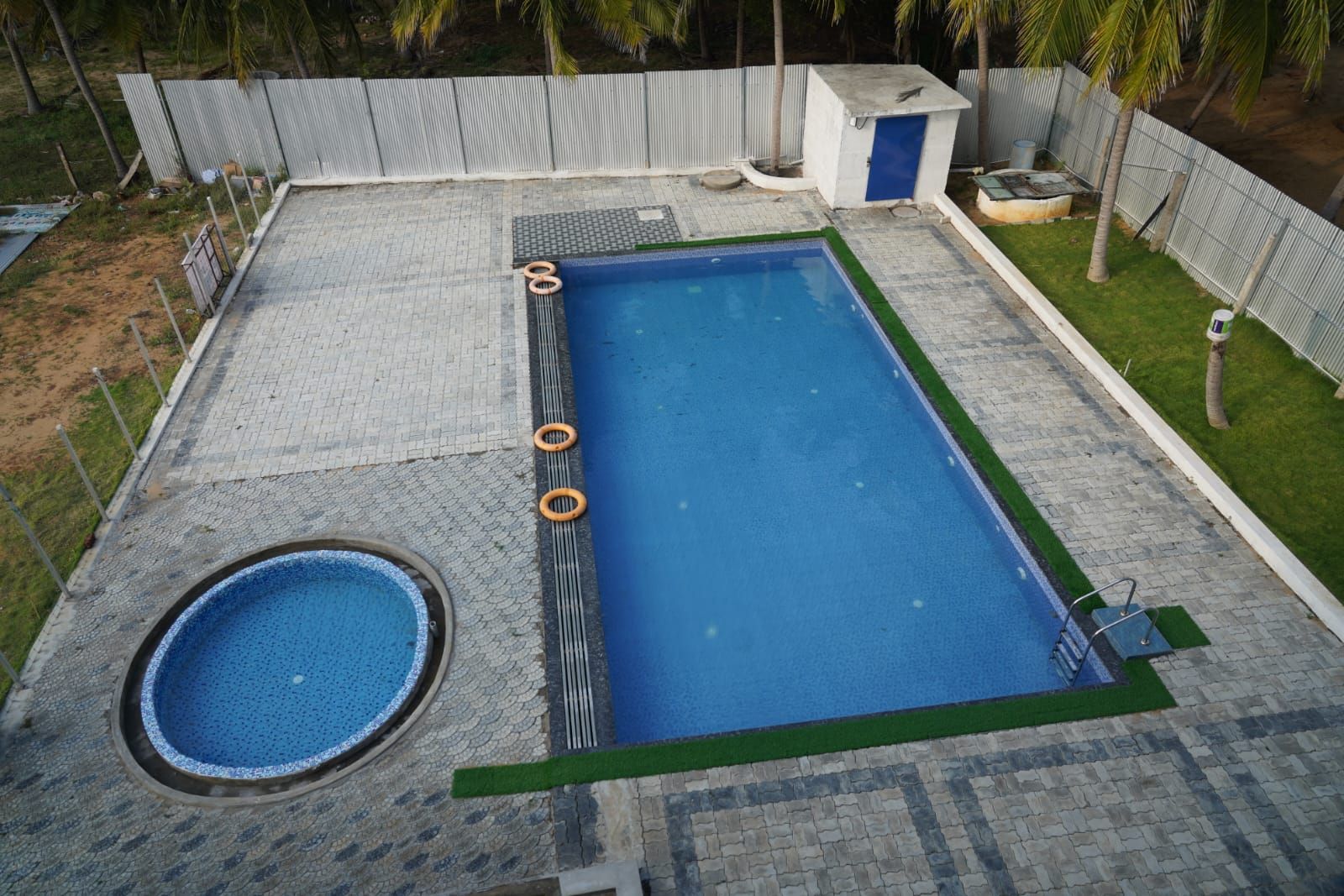 A rectangular swimming pool with a small circular hot tub, surrounded by a paved deck and a metal fence.