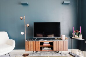 Ein modernes Wohnzimmer mit einem Flachbildfernseher auf einer Holzkonsole, einem weißen Sessel und einem kleinen Beistelltisch mit Kerzen und einer Parfumflasche.