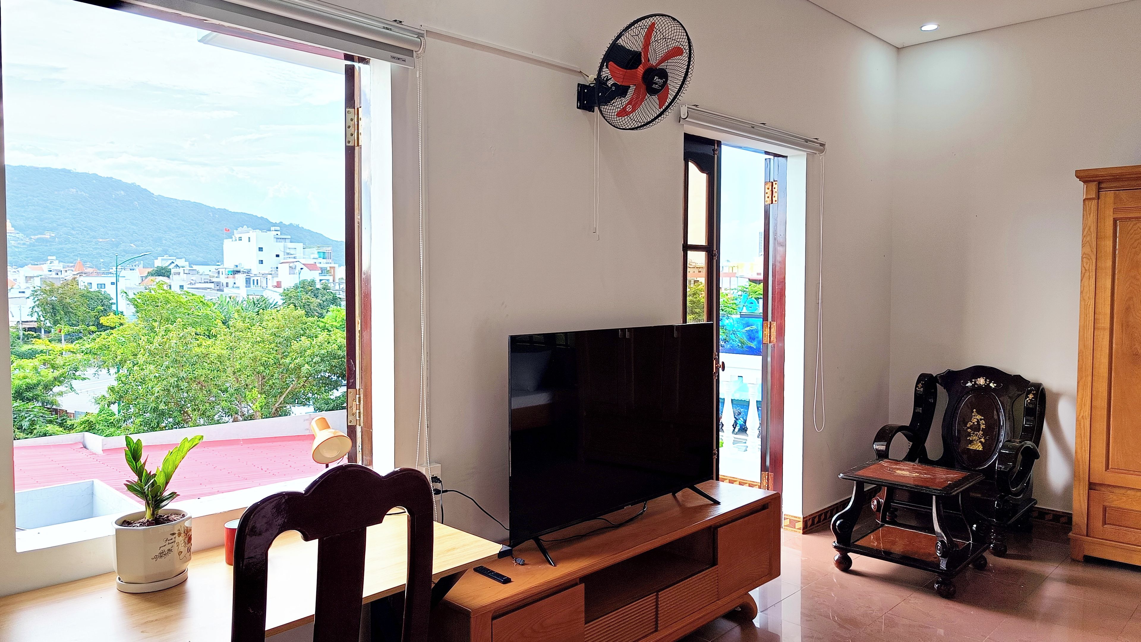 Una sala de estar con televisor sobre un mueble de madera, una silla y un amplio ventanal con vistas a edificios y zonas verdes.