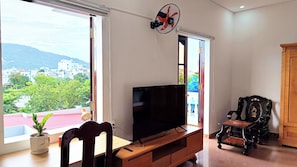 Ein Wohnzimmer mit Fernseher auf Holzfuß, einem Sessel und einem großen Fenster mit Blick auf Gebäude und Grünflächen.