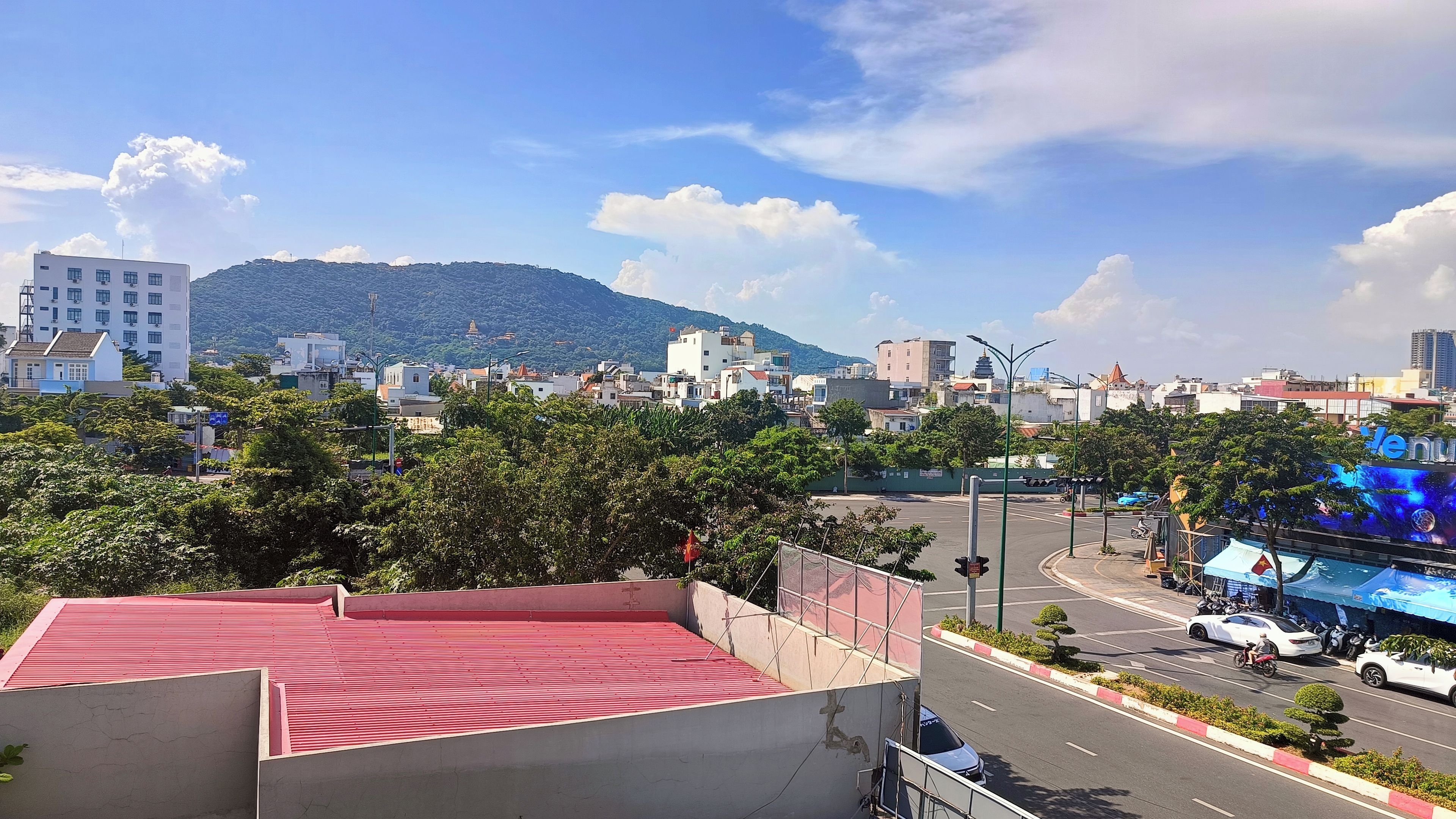 Un paisaje urbano con una colina destacada al fondo, una carretera con vehículos, y un edificio con techo rojo en primer plano.