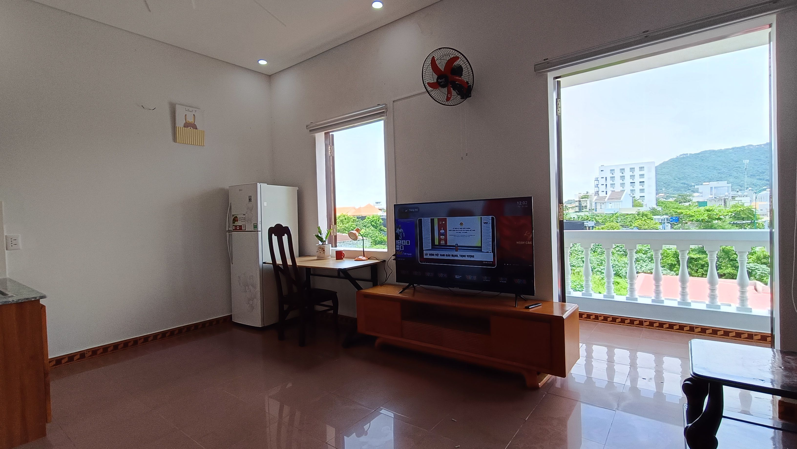 Un espacio habitable compacto con kitchenette, televisor y balcón con vistas a edificios y áreas verdes.