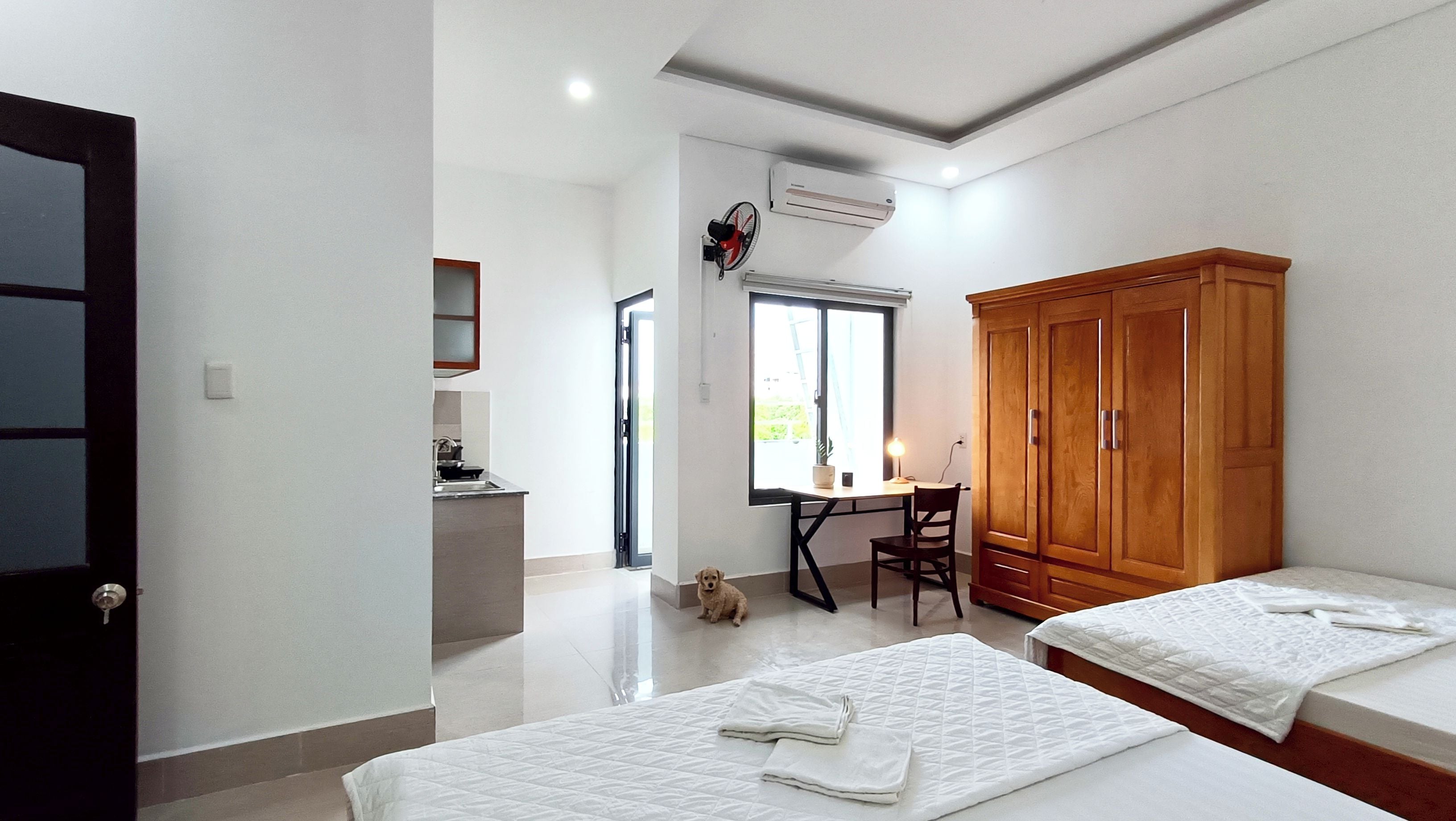 A room with two beds, a desk with a chair, a wardrobe, and a small kitchen area.