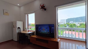 Ein Zimmer mit Fernseher auf einem Schrank, einem Schreibtisch mit Stuhl, einem kleinen Kühlschrank und einem Fenster mit Blick auf Gebäude und Grünflächen.