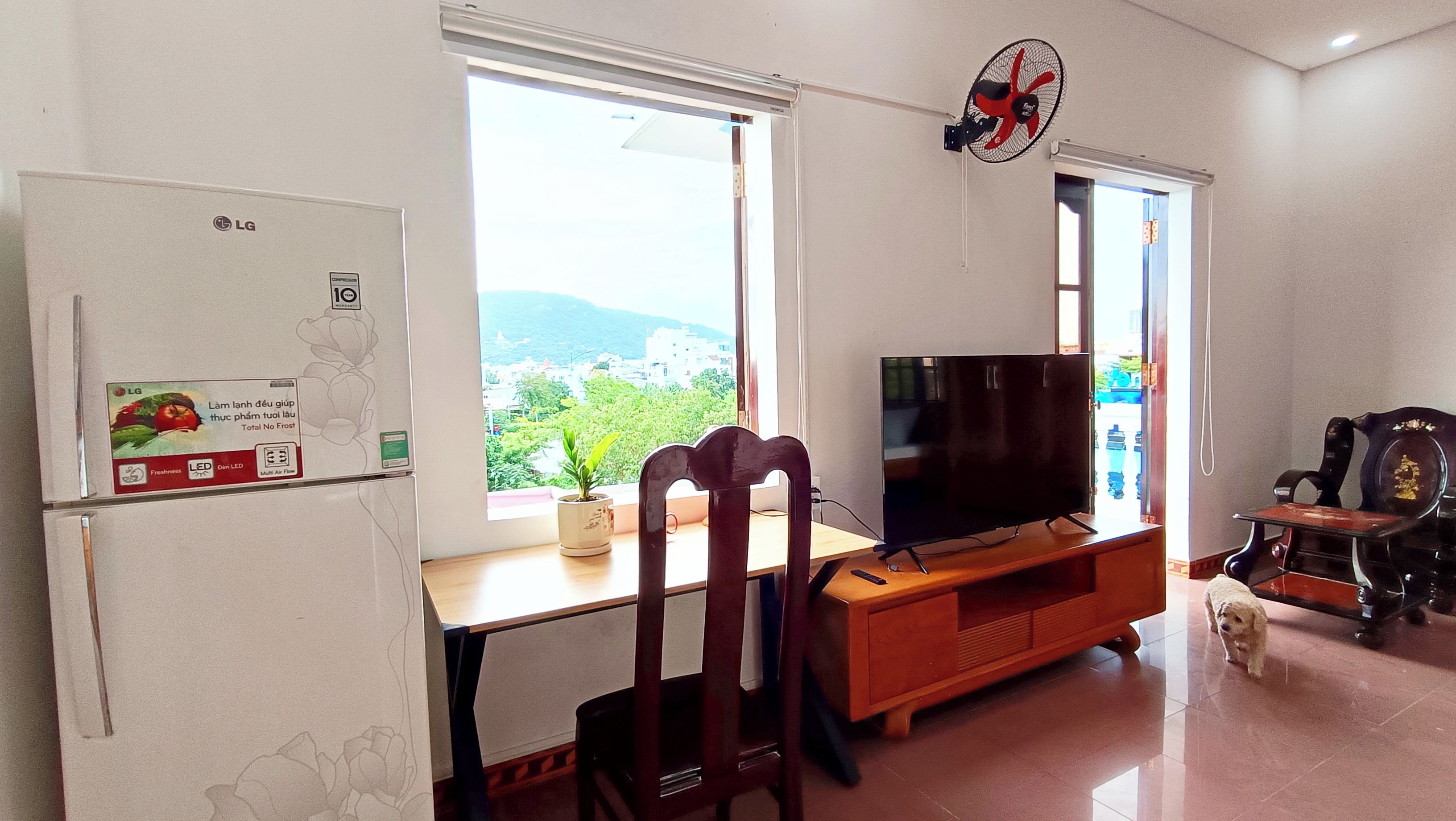 A living room with a TV, a refrigerator, a wooden table, a chair, a small plant, and a dog.