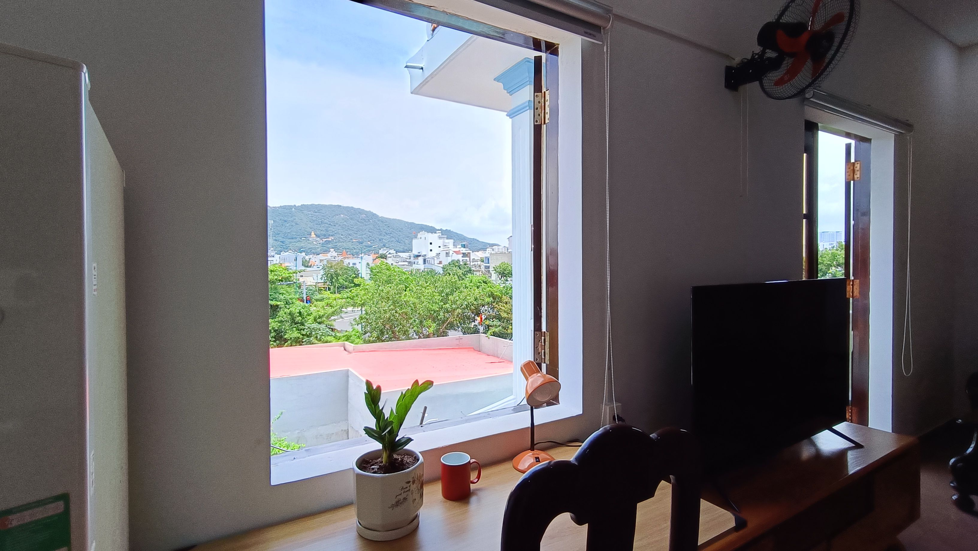 Una habitación con un ventanal grande, un televisor sobre un mueble, una planta en maceta y una taza roja en el alféizar.