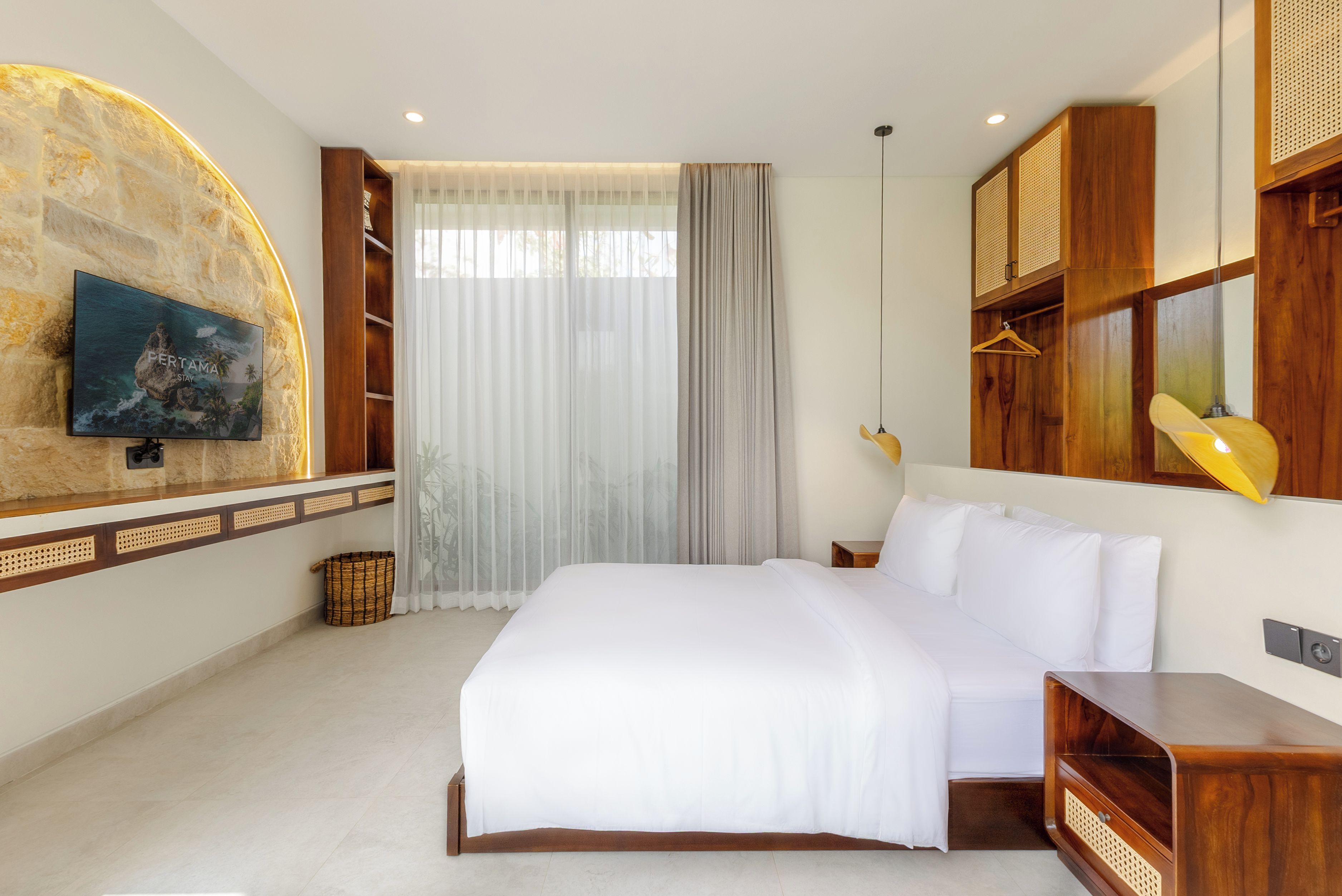 Habitación de hotel con una cama, televisor montado en la pared, ventana con cortinas translúcidas y muebles de madera.