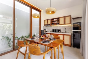 A modern kitchen with wooden cabinets, a dining table set for four, and a large window overlooking a balcony. - Ayuna Villa Uluwatu by Pertama Stay (Badung Regency)