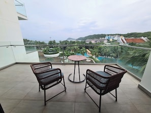 A balcony with two wicker chairs and a small round table, overlooking a pool area with palm trees and a view of a coastal landscape. - Lagoon Residences (Phu Quoc)