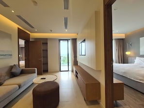 A modern hotel room with a sofa, a small ottoman, a bench, a bed, and a sliding door leading to a balcony. - Lagoon Residences (Phu Quoc)