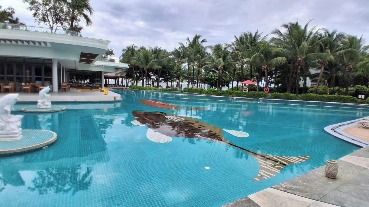 Piscina com escultura de golfinho, palmeiras e um edifício com área de estar ao ar livre.