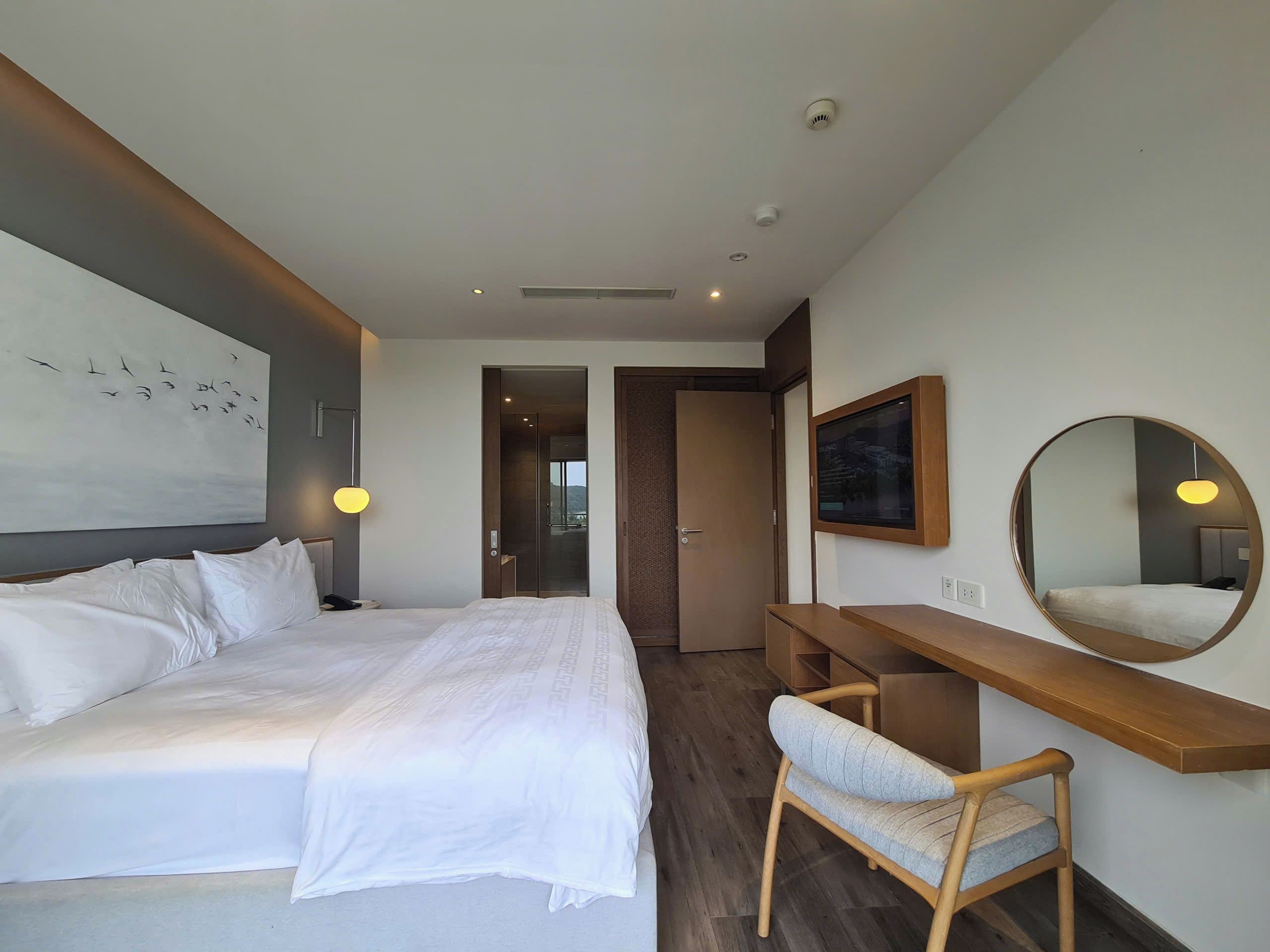Quarto de hotel com cama, escrivaninha, cadeira, espelho e um quadro na parede.