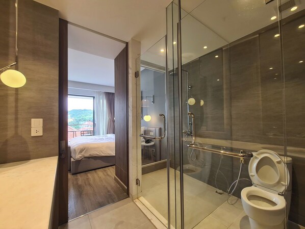 A modern bathroom with a glass-enclosed shower, a toilet, and a visible bed in the adjacent room. - Lagoon Residences  (Phu Quoc)