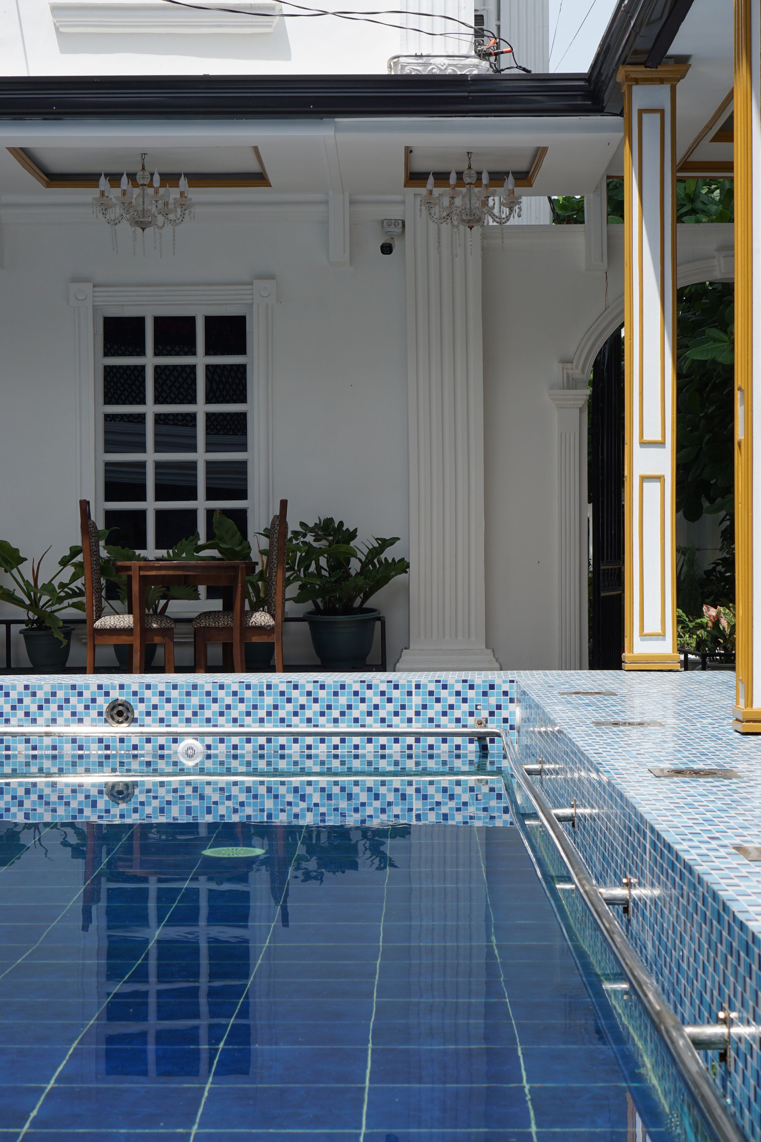 En swimmingpool med blå mosaikplattor, en trästol och krukväxter.
