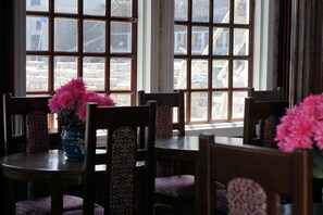 A dining area with a table and chairs, a vase with flowers, and a view through large windows.