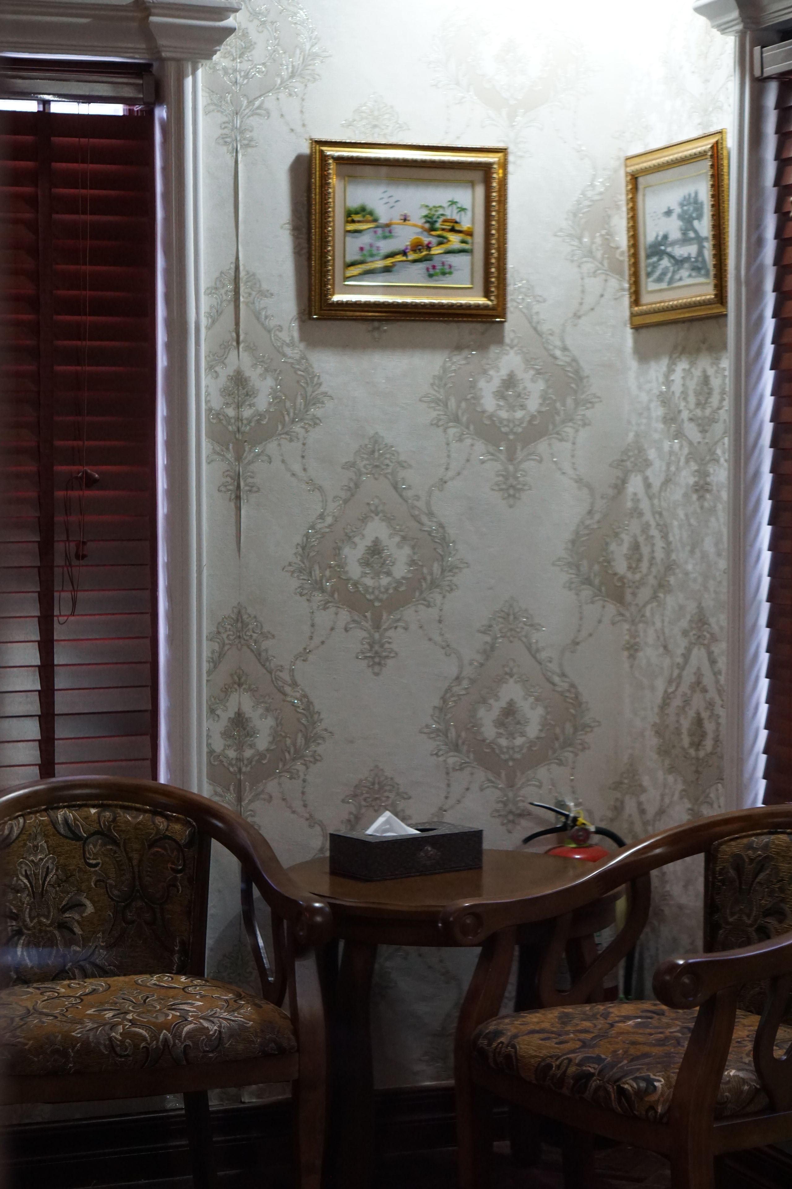 Una habitación con papel tapiz estampado, dos cuadros en la pared, una mesa con una caja de pañuelos y dos sillones tapizados.