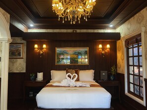 A hotel room with a large bed, a chandelier, wall-mounted lamps, a painting, and a window with a view.