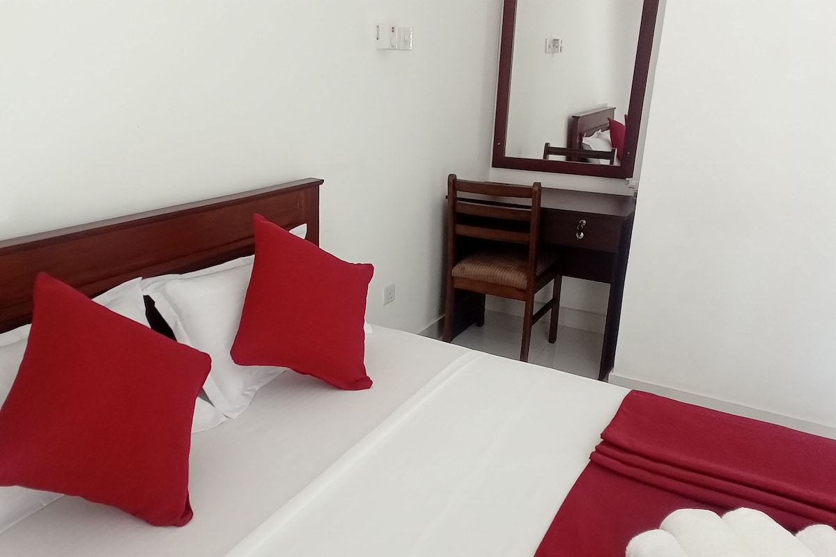 Habitación de hotel con una cama, dos almohadas rojas, un cabecero de madera, un escritorio con una silla y un espejo.