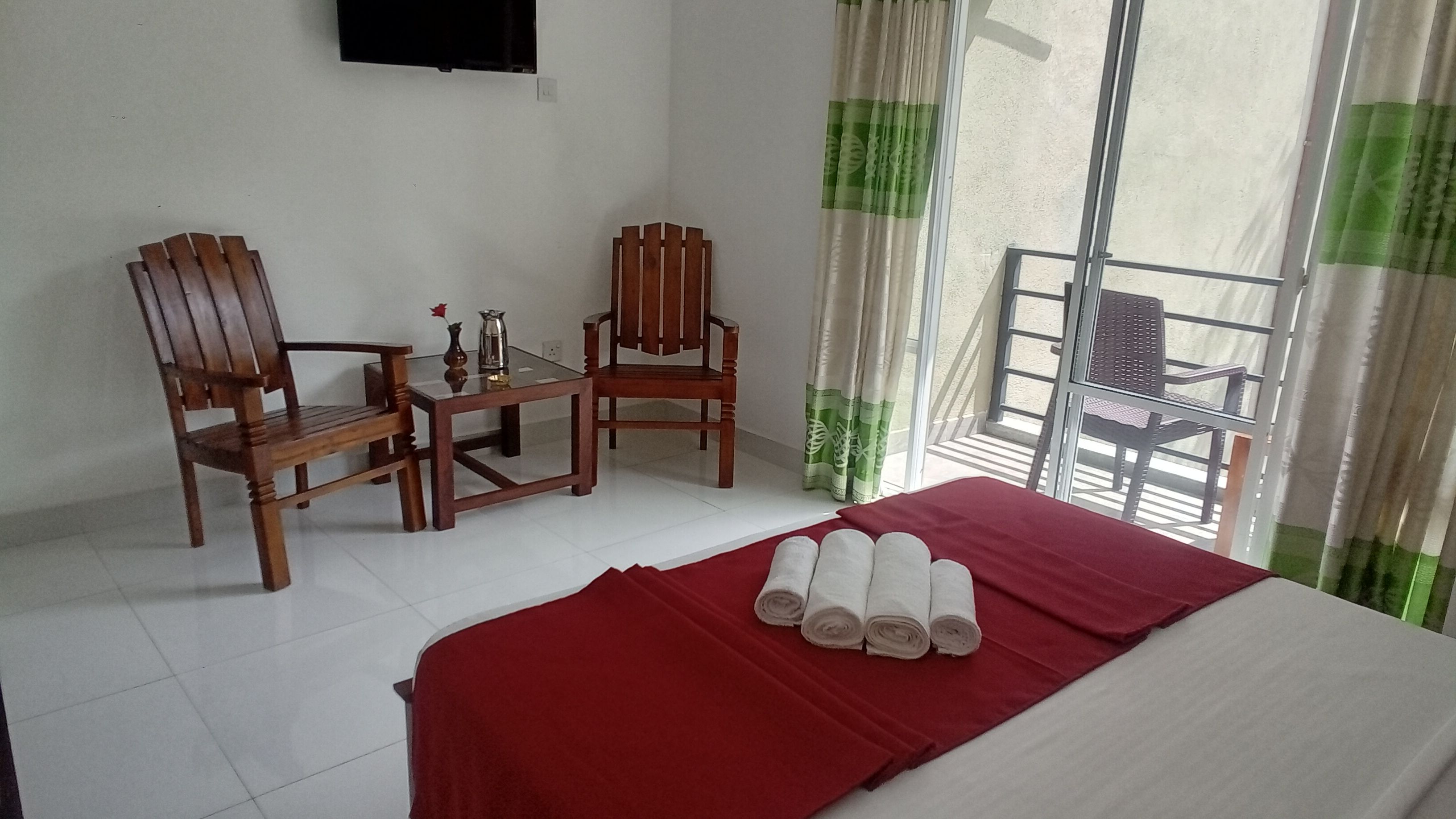 Habitación de hotel con cama, dos sillas de madera, una mesita con un florero y una jarra, y un balcón con una silla.